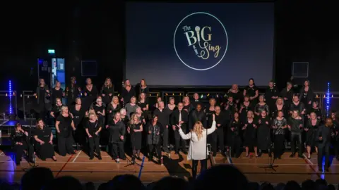 The BIG Sing A large choir dressed in black clothing performing for an audience in front of the BIG Sing logo. A woman in white is conducting the choir.