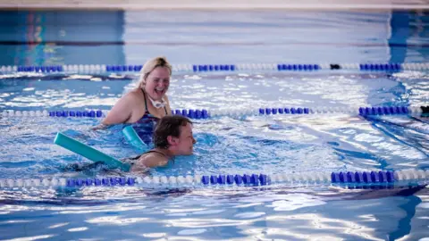 Wimborne Wagtails Two women in a swimming pool, one is stood with goggles around her neck and laughing while another is swimming beside her while using a noodle shaped float.