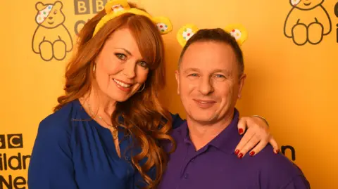 
Gabrielle Blackman (L) and Mark Millar of DIY SOS backstage at BBC Children in Need's 2019 Appeal night at Elstree Studios. Mark is wearing a purple shirt and Gabrille is wearing a navy dress, they both have Pudsey's ear headband on and are staying in front of Pudsey bear and BBC Children in Need logo on a yellow wall.
