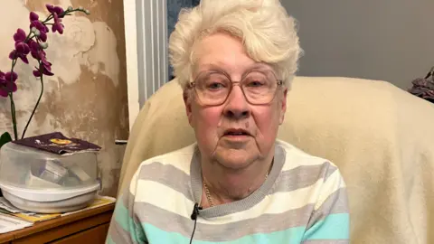 BBC Wendy Hinton, a woman in her early 70s, looks into the camera. She has short white-grey hair, wears a striped top and thin metal-rimmed glasses. Behind her chair you can see a water-damaged wall, an orchid and an interior door that leads to another room.