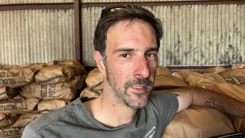 Luke Ablett, 36, has dark hair swept across his head and a stubbly beard and moustache with some grey flecks in, he is wearing a grey t-shirt, behind him are stacks of 25kg bags of potatoes. 