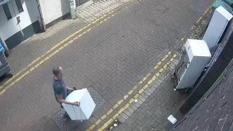 CCTV image of a man on a small street carrying a small white fridge in the direction of where two other fridges have been left on the side of the road.