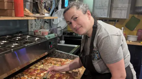 Suzy Sales in the kitchen wearing an apron and leaning over some pizzas as she continues to prepare them