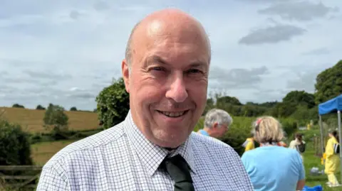A man with a bald head and a white checked shirt and green tie on.