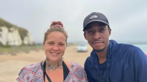 A man and a woman stand together next to a beach. They are smiling. 