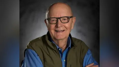 Robert Lawrence Merhaut A man with no hair and dark glasses is smiling at the camera. He is wearing a blue shirt and a green gilet. 