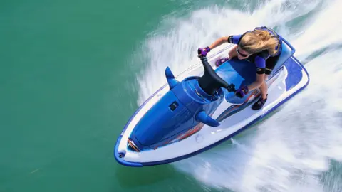 A jet ski rider tears through the water with ripples appearing either side of her in the water. She is wearing protective gloves and glasses.