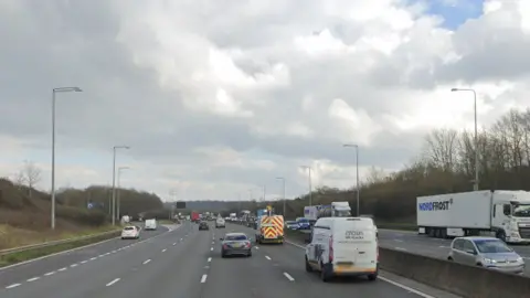 A general view of the M25 clockwise carriageway close to Waltham Abbey. Three lanes of traffic can be seen as well as a slip road leading to the town. Vehicles can be seen heading along the road including on the opposite carriageway.