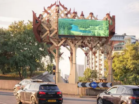Wildstone Estates Ltd/Tate + Co CGI image of advertising board on large beige and brown structure with cars in foreground on roundabout