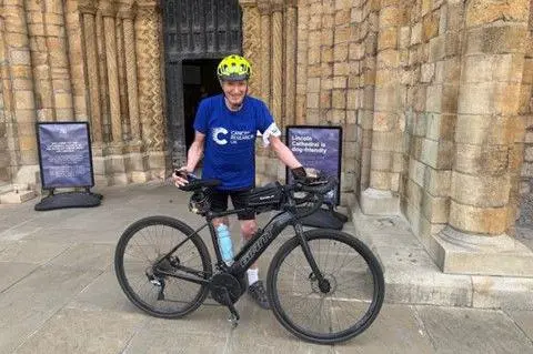 John Whitbread A man in cycling gear standing in front of the entrance to Lincoln Cathedral. He is wearing a blue T-shirt and is pictured with his black-coloured bike.