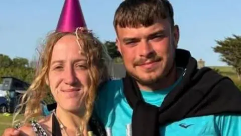 Jamie Chappell, a young man with dark hair and a beard, is standing in a field wearing a party hat next to a young woman holding a cake.