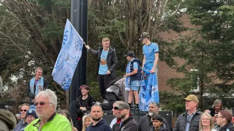 Fans standing on make-do platforms to get a better view. One is carrying a Coventry City flag