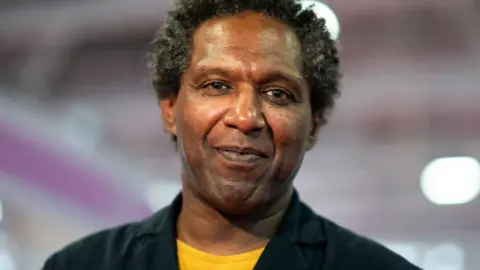 Getty Images Lemn Sissay, a man in a yellow t-shirt and black jacket, standing against a purple backdrop. 