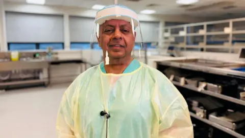 An image of Paul Bachoo in surgical protective wear, smiling at camera, in a hospital setting.