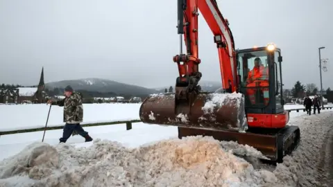 An orange digger clearing deep snow in Aboyne in Aberdeenshire as people walk past.