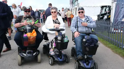 Matt Keeble/PA Wire Two men and a woman at the Royal Cornwall Show. They are on mobility scooters and looking directly at the camera as people walk past them. The mobility scooters have baskets on the front, which are filled with flasks, water bottles, jumpers and bags. 