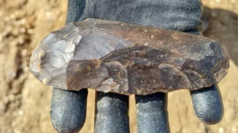 A Neolithic polished axe held out on a gloved hand, clad in black. Beneath it is sandy soil. 