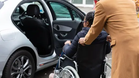 A parent helping their child who is in a wheelchair getting into a silver car.
