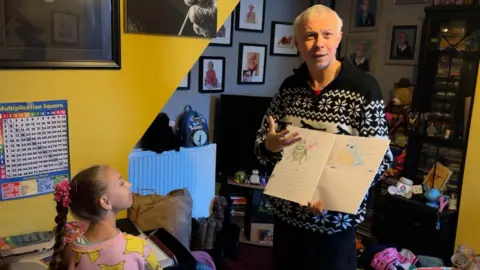 BBC Clarke is being watched by daughter Dakota as he holds open an exercise book containing colourful pictures of Pokemon characters. 