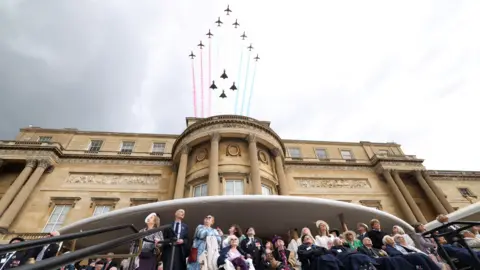 Red Arrows deploy coloured smoke as they fly over Buckingham Palace with a group of veterans all looking up to the sky below. It's a moody, grey cloudy day.