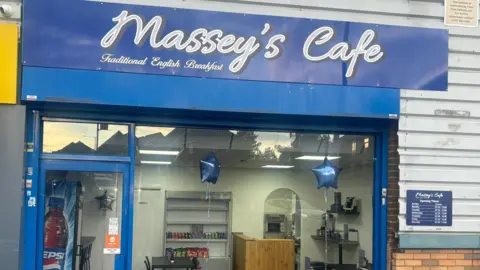 The window front of a blue cafe named Massey's Cafe. It has blue star balloons in the window and a black table can be seen inside. There is a brown counter top in the far right corner.