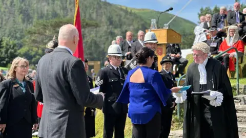 A woman in a blue top with flowing sleeves hands a petition in a blue plastic pocket to the Clerk of Tynwald, who is wearing a black robe and white wig. A man in a grey suit waits behind with his document. People in formal wear, including police in formal dress, line the area at the foot of Tynwald Hill.