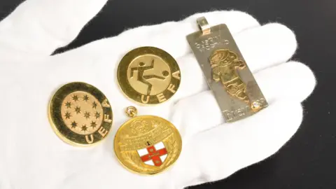 Mullock Jones Auctioneers & Valuers Four medals on a white gloved hand. Three are circular and gold, with two that say "UEFA", and one with a red and white St George's cross on it. The fourth is rectangular and silver, with a gold man on it, kicking a football.