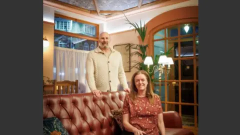 CARE charity Man with a beard who is wearing a cream cardigan and is standing behind a brown leather sofa. A woman is sitting on the sofa and she is smiling and has long brown hair and is wearing a brown patterned top. The walls are a peach colour at the bottom and cream on top and the ceiling is patterned with a blue William Morris design. There is a window with a cafe style voile curtain and there is a standard lamp with 3 white shades. There is a tall green plant and a picture on the wall.