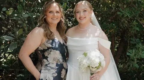 Kylee Lynn Photography Hannah and her mother at Hannah's wedding. Her mother is wearing a blue dress with white flowers. Hannah on the right is dressed in a white wedding dress holding a bouquet