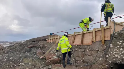 Government of Jersey A group of workers working on a damaged seawall. There is a wooden structure around the wall with a two men working on it. In front of the wall is rocks covered in seaweed and shells. 