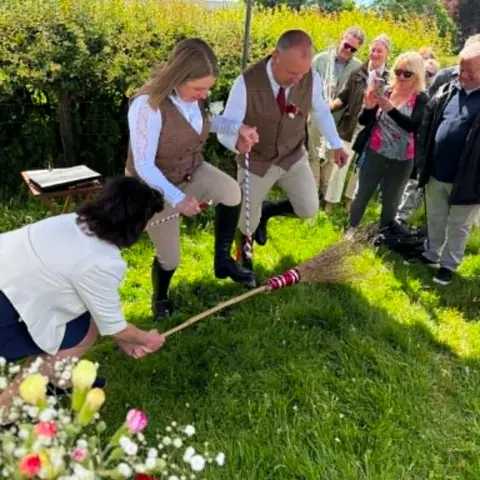 Family photo Safron and Nigel dancing under a black marquee with friends and family watching them. They are dancing over a brown broom stick which a woman holds under their legs. 