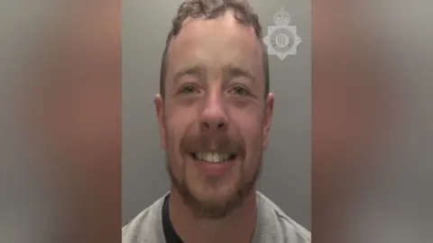 A police custody image of man with short brown hair and a beard smiling into the camera. He is wearing a grey sweatshirt and is sitting in front of a plain background.