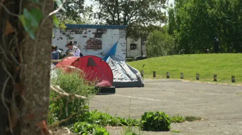 A grey and red tent in a park with homeless outreach workers in the background speaking to asylum seekers to support them