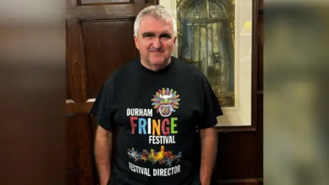 Handout Stephen Cronin standing in front of a picture, wearing a Durham Fringe T-shirt 