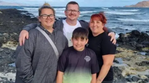 John (middle) stands with his family Molly (left) Amanda (right) and Charlie (bottom middle) on a beach. He has his arms around Molly and Amanda and they are all smiling at the camera.