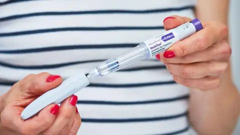 A woman in a blue and white stripped t-shirt holds a Mounjaro injection pen.