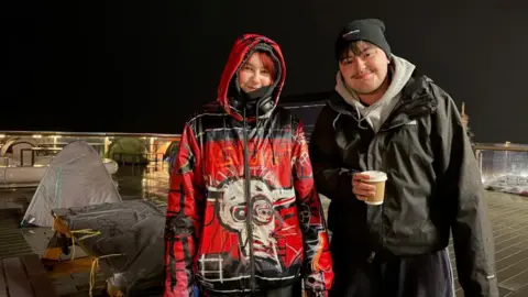 Amber and Noah pose for a photograph, smiling to the camera with their backs to the Brighton seafront. The sky is pitch black. The ground is wet, showing signs of heavy rain. Both are wearing waterproof jackets. Amber has her hood up with over-ear headphones hanging around her neck. Noah is wearing a beanie while holding a coffee cup in his right hand. A bin bag is spread out across the top of a bench directly behind the pair. Tents are propped up and visible in the background, further along the deck at the base of the i360.