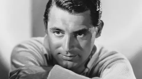 Cary Grant posing in front of a camera in a black and white photograph. He has neatly combed hair, an intense, thoughtful expression, with his chin resting on his folded arms. He is wearing a textured sweater over a shirt. 