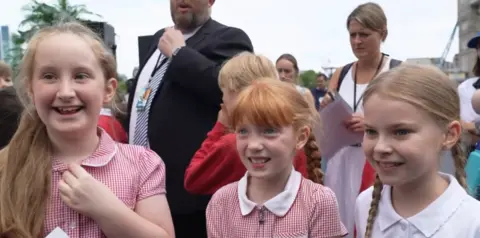 Three girls wearing red and white school uniforms. They are standing next to each other and looking to the side of the camera. They are all smiling. 