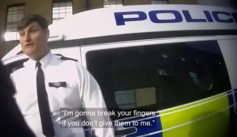 A screenshot from undercover filming showing an officer in a uniform made up of a white shirt and a black tie standing in front of the side of a police vehicle. A subtitle on the image reads "I'm gonna break your fingers if you don't give them to me."