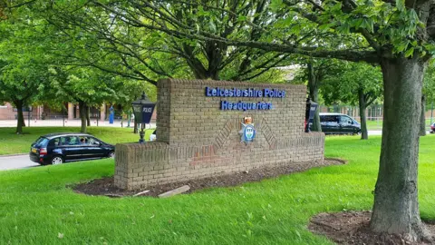 A brick sign for the force HQ with a blue lamp and a crest on it, on a grassy verge