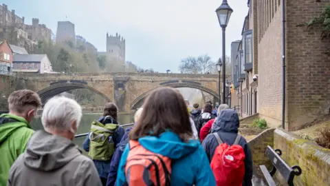 Back view of the head and shoulders of a group of walkers on a path with buildings to one side and a river on the other. They are wearing anoraks or kagoules and some have small backpacks.  Ahead of them an arched stone bridge spans the river and Durham Cathedral and Castle stand to one side above it.