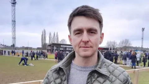 A man with short brown hair wearing a grey jumper and green coat looks at the camera. Behind him fans and footballers are standing pitch side. 