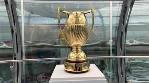 Sam Dixon-French/BBC The Women's Rugby World Cup trophy - gold coloured with two large handles and ornate carvings. It is standing on a plinth in front of curved glass windows.