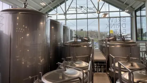 Alex Pope/BBC The inside of a brewery, showing large silver metal containers, and a walkway. There is a large glass window with writing on. The ceiling is white. 
