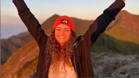 Jamie Graham Maria Eftimova standing in the mountains, wearing a red hat and fleece, smiling at the camera and raising her hands in the air above her head.