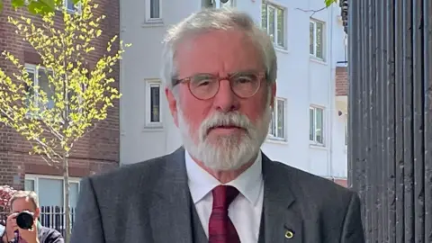 Gerry Adams is pictured from the chest up - he wears a grey jacket, grey waistcoat and red tie. He has two pins on his lapel. He has grey hair, a grey beard and brown glasses. In the background you can see a photographer taking a picture. 