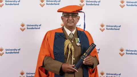Ben Banerjee receiving his honorary doctorate of medicine from the University of Sunderland. He is wearing an orange academic cap and gown as well as a brown military uniform. He is holding a scroll and looking into the camera.