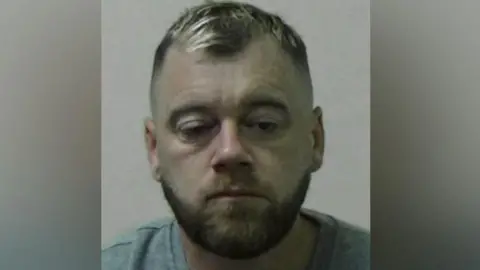 Mugshot of Steven Rycroft. He has short dark hair with blond highlights and a brown beard. His right eye is partially closed and he looks like he is dazed. He is wearing a grey shirt.