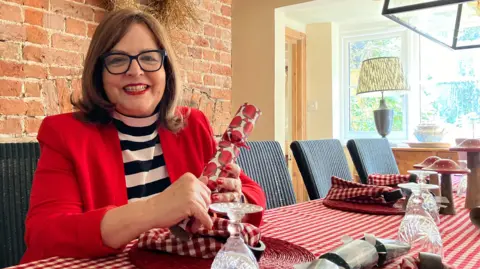 Lewis Adams/BBC Jane Malyon sat at the table holding a cracker. The table cloth has red and white checkers and has napkins, crackers and glasses on it. Mrs Malyon herself is smiling at the camera.